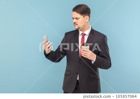 Portrait of serious concentrated man with mustache standing using mobile phone and drinking takeaway coffee, wearing black suit with red tie. Indoor studio shot isolated on light blue background. 105857064