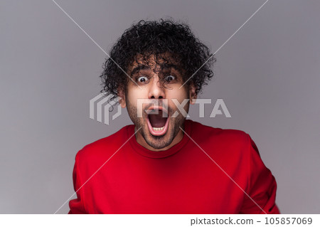 Portrait of amazed astonished surprised bearded man with curly hair, looking at camera with big eyes and open mouth, wearing red jumper. Indoor studio shot isolated on gray background. Portrait of amazed astonished surprised bearded man with curly hair, looking at camera with big eyes and open mouth, wearing red jumper. Indoor studio shot isolated on gray background. 105857069