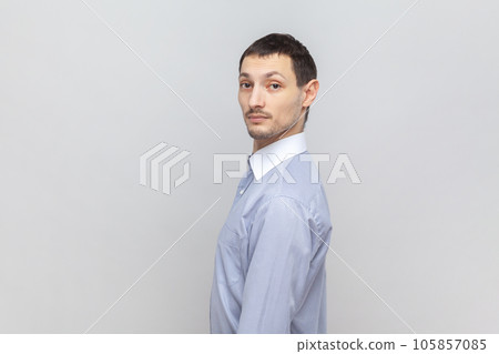 Side view portrait of serious attractive man standing looking at camera with strict concentrated facial expression, wearing light blue shirt. Indoor studio shot isolated on gray background. Side view portrait of serious attractive man standing looking at camera with strict concentrated facial expression, wearing light blue shirt. Indoor studio shot isolated on gray background. 105857085