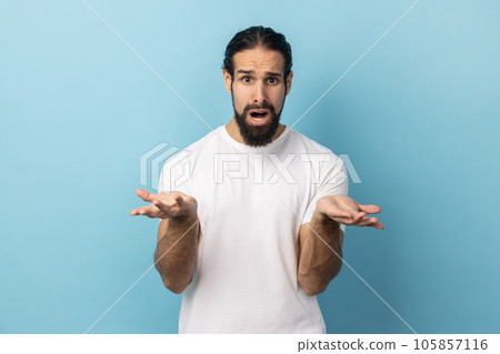 What do you want. Confused man with beard wearing white T-shirt standing with raised hands and surprised indignant expression, asking what reason. Indoor studio shot isolated on blue background. 105857116