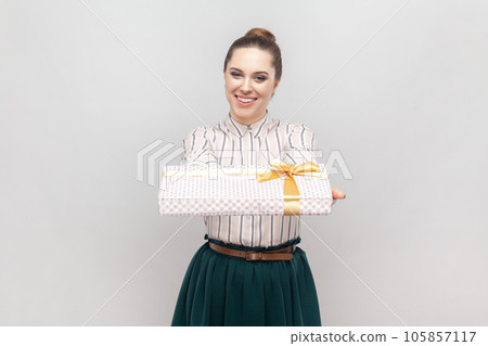 Portrait of pleased pretty attractive woman wearing striped shirt and green skirt standing with gift box in hand, giving present, congratulating. Indoor studio shot isolated on gray background. 105857117
