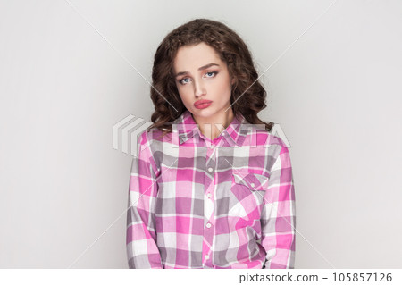 Portrait of sad lonely woman purses lips, feels bored looks uninterested and tired, listens boring stories from friend, wearing pink checkered shirt. Indoor studio shot isolated on gray background. Portrait of sad lonely woman purses lips, feels bored looks uninterested and tired, listens boring stories from friend, wearing pink checkered shirt. Indoor studio shot isolated on gray background. 105857126
