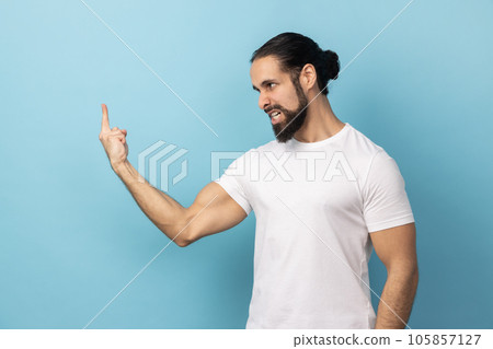 Side view of man with beard wearing white T-shirt showing middle finger, rude gesture, aggressive protest with impolite finger sign, expressing hatred. Indoor studio shot isolated on blue background. 105857127