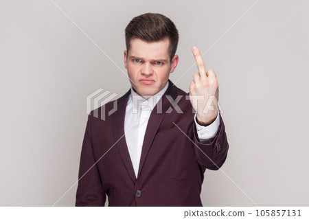 Portrait of man shows fuck off sign being rude and looks dissatisfied at camera, expresses bad attitude to someone, wearing violet suit and white shirt. Indoor studio shot isolated on grey background. 105857131