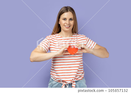 Portrait of smiling adorable blond woman wearing striped T-shirt holding little red heart in front her chest, expressing romantic feeling and love. Indoor studio shot isolated on purple background. 105857132