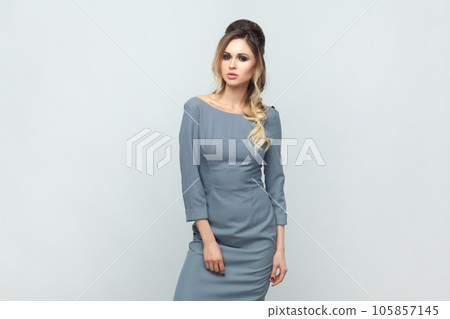 Confident attractive woman fashion model with hairstyle and bright makeup, standing looking at camera with serious face, wearing grey elegant dress. Indoor studio shot isolated on gray background. 105857145
