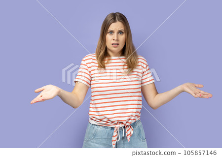 I don't know. Blond woman wearing striped T-shirt standing with wide raised arms and looking at camera with frowning face, and dont know what to do. Indoor studio shot isolated on purple background. I don't know. Blond woman wearing striped T-shirt standing with wide raised arms and looking at camera with frowning face, and dont know what to do. Indoor studio shot isolated on purple background. 105857146