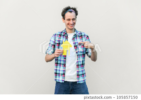 Portrait of nice attractive glad cheerful man holding in hands paper house form, sale bank loan purchase, wearing blue checkered shirt and headband. Indoor studio shot isolated on gray background. 105857150