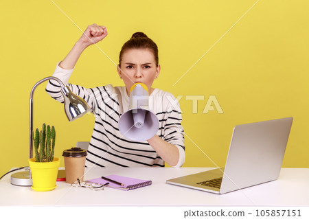 Portrait of angry woman loudly screaming at megaphone, making announce, protesting, wants to be heard, sitting on workplace. Indoor studio studio shot isolated on yellow background. Portrait of angry woman loudly screaming at megaphone, making announce, protesting, wants to be heard, sitting on workplace. Indoor studio studio shot isolated on yellow background. 105857151