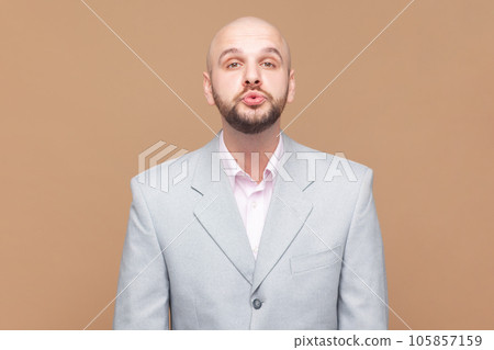 Portrait of bald bearded man keeps lips rounded, blows air mwah, expresses love to girlfriend, shows passion and affection, wearing gray jacket. Indoor studio shot isolated on brown background. 105857159