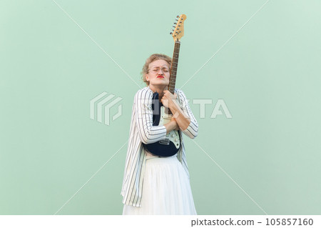 Portrait of blonde woman in white shirt, skirt, and striped blouse with eyeglasses standing with closed eyes and embracing guitar. Indoor studio shot isolated on light green background. Portrait of blonde woman in white shirt, skirt, and striped blouse with eyeglasses standing with closed eyes and embracing guitar. Indoor studio shot isolated on light green background. 105857160