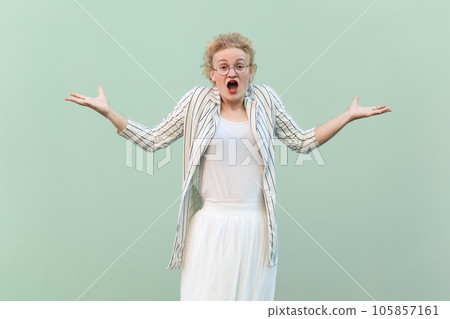 Portrait of shocked confused blonde woman wearing striped shirt and skirt, spread hands, asking what, looking at camera, screaming. Indoor studio shot isolated on light green background. Portrait of shocked confused blonde woman wearing striped shirt and skirt, spread hands, asking what, looking at camera, screaming. Indoor studio shot isolated on light green background. 105857161