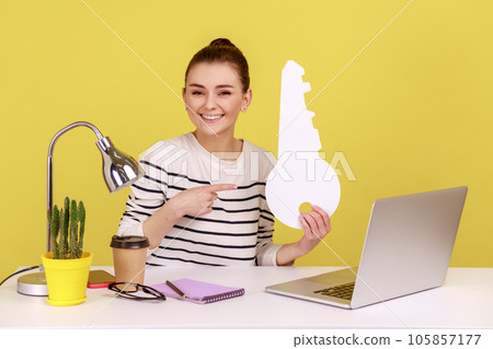 Amazing woman sitting at workplace with laptop, pointing at big paper key and smiling, satisfied with new home, rental office. Indoor studio studio shot isolated on yellow background. Amazing woman sitting at workplace with laptop, pointing at big paper key and smiling, satisfied with new home, rental office. Indoor studio studio shot isolated on yellow background. 105857177