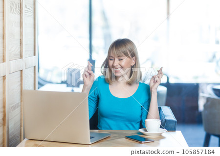 Portrait of happy positive hopeful young woman with blonde hair in blue shirt working on laptop, crossing fingers, making wish, keeps eyes closed. Indoor shot in cafe with big window on background. 105857184