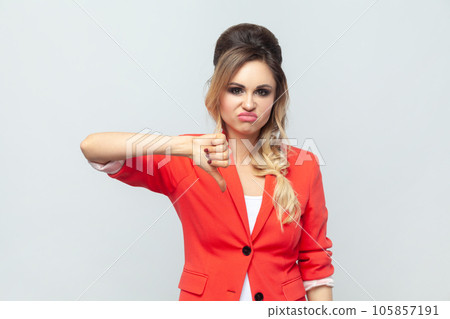 Portrait of disappointed attractive upset sad blonde woman standing showing thumb down, dislike gesture, disagree, wearing red jacket. Indoor studio shot isolated on gray background. 105857191