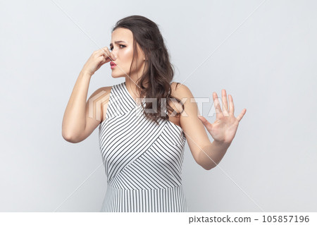 Displeased woman covers nose with hand, smells something awful, pinches nose, frowns in displeasure, sees pile of garbage, wearing striped dress. Indoor studio shot isolated on gray background. 105857196