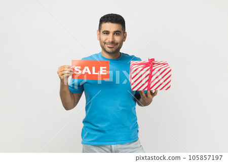Portrait of positive optimistic unshaven man wearing blue T- shirt standing with present gift box and sale card, discounts for presents before holidays. Indoor studio shot isolated on gray background. 105857197