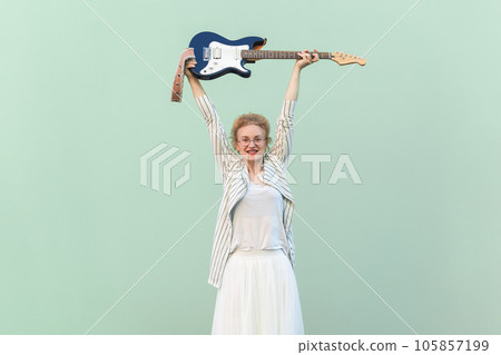 Portrait of joyful cheerful blonde woman in striped shirt and white skirt with eyeglasses raised her arms and holding guitar. Indoor studio shot isolated on light green background. Portrait of joyful cheerful blonde woman in striped shirt and white skirt with eyeglasses raised her arms and holding guitar. Indoor studio shot isolated on light green background. 105857199