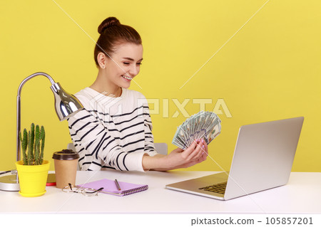 Satisfied rich woman office manager showing hundred dollar bills to laptop display, sitting at workplace, satisfied about big profit. Indoor studio studio shot isolated on yellow background. 105857201