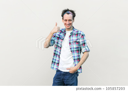 Portrait of man in casual blue checkered shirt and headband approves incredible promo, keeps thumbs up likes and agrees being satisfied with something. Indoor studio shot isolated on gray background. Portrait of man in casual blue checkered shirt and headband approves incredible promo, keeps thumbs up likes and agrees being satisfied with something. Indoor studio shot isolated on gray background. 105857203