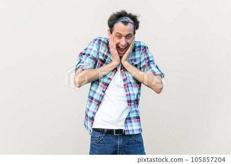Wow, that great. Overjoyed handsome man keeps hands on cheeks, looks joyfully at camera, being amazed, wearing blue checkered shirt and headband. Indoor studio shot isolated on gray background. Wow, that great. Overjoyed handsome man keeps hands on cheeks, looks joyfully at camera, being amazed, wearing blue checkered shirt and headband. Indoor studio shot isolated on gray background. 105857204
