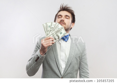 Portrait of rich delighted proud bearded man standing smelling dollar banknotes, being satisfied of big salary, wearing grey suit and blue bow tie. Indoor studio shot isolated on gray background. Portrait of rich delighted proud bearded man standing smelling dollar banknotes, being satisfied of big salary, wearing grey suit and blue bow tie. Indoor studio shot isolated on gray background. 105857205
