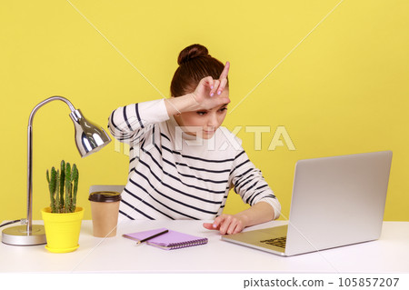 Lost job, unemployment. Depressed woman sitting at workplace with laptop and showing loser gesture, desperate about unlucky dismissal. Indoor studio studio shot isolated on yellow background. Lost job, unemployment. Depressed woman sitting at workplace with laptop and showing loser gesture, desperate about unlucky dismissal. Indoor studio studio shot isolated on yellow background. 105857207