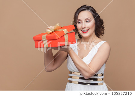 Portrait of smiling optimistic middle aged woman with wavy hair opens present box, looking inside, being interesting what inside, wearing white dress. Indoor studio shot isolated on light brown 105857210