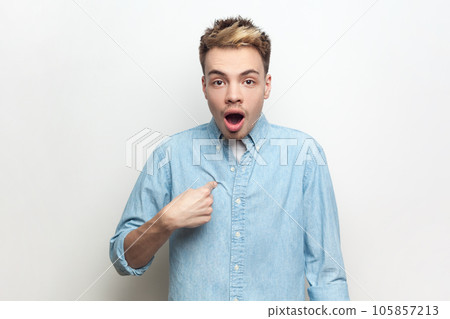 Portrait of shocked astonished surprised man wearing denim shirt pointing finger at himself, looking at camera with big eyes and open mouth. Indoor studio shot isolated on gray background. 105857213