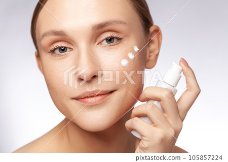 Closeup portrait of adorable well groomed beautiful woman holding bottle with lotion applying cream on her face, beauty routine. Indoor studio shot isolated over gray background. 105857224