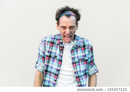 Naughty comic man sticks out tongue, misbehaves, demonstrates her disobedience and protest, has funny look, wearing blue checkered shirt and headband. Indoor studio shot isolated on gray background. 105857228