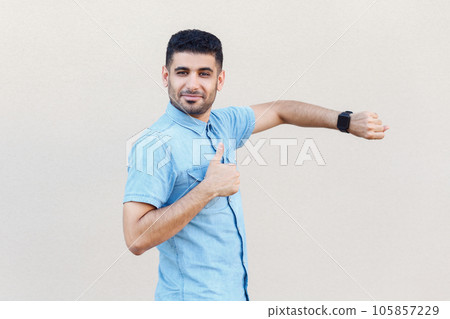 Portrait of positive attractive calm man wearing denim shirt standing showing his smart watch and thumb up, like gesture, recommend. Indoor studio shot isolated on gray background. Portrait of positive attractive calm man wearing denim shirt standing showing his smart watch and thumb up, like gesture, recommend. Indoor studio shot isolated on gray background. 105857229