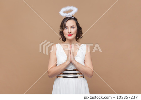 Portrait of hopeful middle aged woman with wavy hair and nimb over head, praying, pleading about something, wearing white dress. Indoor studio shot isolated on light brown background. 105857237