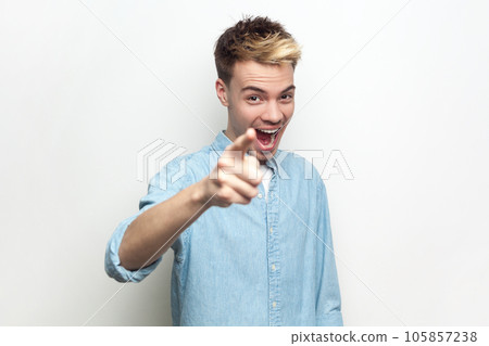 Portrait of excited positive smiling handsome man wearing denim shirt pointing finger to camera, selecting you, being in good mood. Indoor studio shot isolated on gray background. Portrait of excited positive smiling handsome man wearing denim shirt pointing finger to camera, selecting you, being in good mood. Indoor studio shot isolated on gray background. 105857238