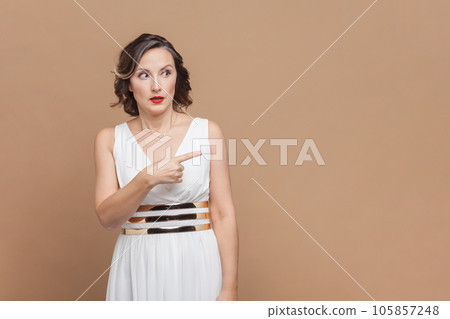 Portrait of shocked surprised middle aged woman with wavy hair pointing away at copy space for advertisement, wearing white dress. Indoor studio shot isolated on light brown background. Portrait of shocked surprised middle aged woman with wavy hair pointing away at copy space for advertisement, wearing white dress. Indoor studio shot isolated on light brown background. 105857248