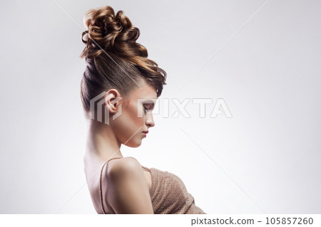 Side view portrait of beautiful brunette young woman with stylish hairdo and makeup, looking away, demonstrates her fashionable hairdo. Indoor studio shot isolated on gray background. Side view portrait of beautiful brunette young woman with stylish hairdo and makeup, looking away, demonstrates her fashionable hairdo. Indoor studio shot isolated on gray background. 105857260
