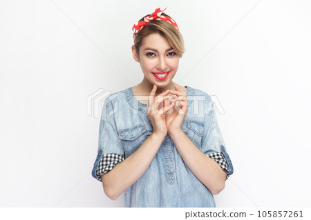 Portrait of sly beautiful blonde woman wearing blue denim shirt and red headband standing looking at camera with bitchy cunning expression. Indoor studio shot isolated on gray background. 105857261