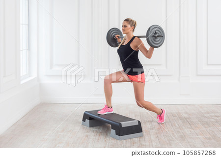 Portrait of sporty beautiful woman wearing pink shorts and black top holding barbell on shoulders and doing squats with one leg on step on white wall background. Indoor studio shot. 105857268