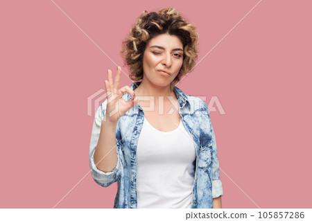 Portrait of playful woman with curly hairstyle wearing blue shirt standing showing okay gesture, winking to camera, saying great job. Indoor studio shot isolated on pink background. 105857286