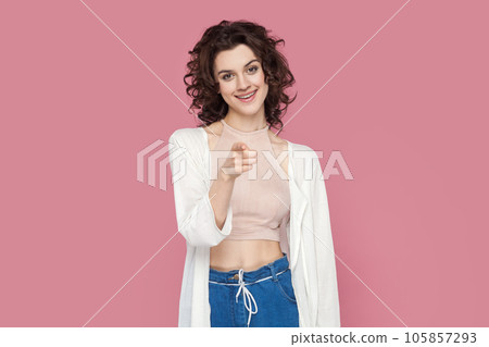 Portrait of satisfied smiling attractive woman with curly hairstyle in casual style outfit pointing at you with finger, expressing positive emotions. Indoor studio shot isolated on pink background. 105857293