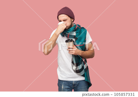 Portrait of unhealthy bearded man in white T-shirt, beany hat and scarf standing holding paper cup and sneezing, catching cold, runny nose. Indoor studio shot isolated on pink background. Portrait of unhealthy bearded man in white T-shirt, beany hat and scarf standing holding paper cup and sneezing, catching cold, runny nose. Indoor studio shot isolated on pink background. 105857331