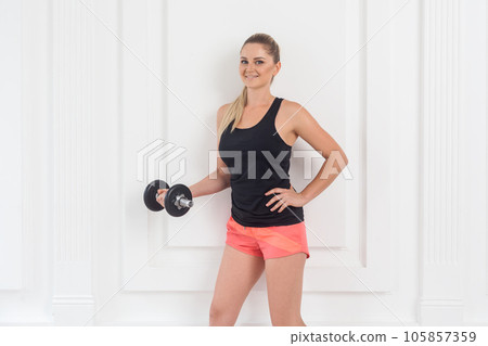 Young smiling athletic beautiful woman in pink shorts and black top holding dumbbells, looking smiling at camera, doing exercise for arms at the gym on white wall background. Indoor studio shot. Young smiling athletic beautiful woman in pink shorts and black top holding dumbbells, looking smiling at camera, doing exercise for arms at the gym on white wall background. Indoor studio shot. 105857359