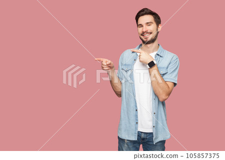 Man in blue casual style shirt standing points away on copy space, discusses amazing promo, give way or direction, has pleasant smile, feels optimistic. Indoor studio shot isolated on pink background Man in blue casual style shirt standing points away on copy space, discusses amazing promo, give way or direction, has pleasant smile, feels optimistic. Indoor studio shot isolated on pink background 105857375
