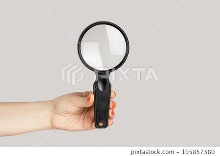 Closeup of woman hand showing loupe, magnificing glass, equipment for inspecting and zooming. Indoor studio shot isolated on gray background. Closeup of woman hand showing loupe, magnificing glass, equipment for inspecting and zooming. Indoor studio shot isolated on gray background. 105857380