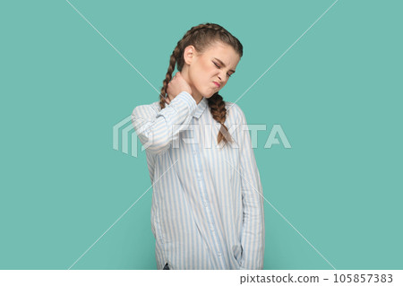 Portrait of teenager girl with braids wearing striped shirt touches neck, suffers osteochandrosis, leads sedentary lifestyle, feels strong pain. Indoor studio shot isolated on green background. 105857383