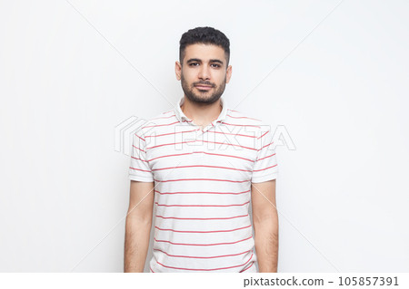 Portrait of calm satisfied delighted bearded man wearing striped t-shirt standing looking at camera, expressing positive emotions. Indoor studio shot isolated on gray background. Portrait of calm satisfied delighted bearded man wearing striped t-shirt standing looking at camera, expressing positive emotions. Indoor studio shot isolated on gray background. 105857391
