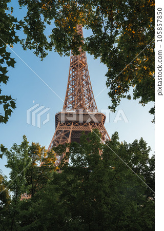Sunny side view of the Eiffel Tower in Paris, France, seen through lush green trees under a clear blue sky. Sunny side view of the Eiffel Tower in Paris, France, seen through lush green trees under a clear blue sky. 105857850