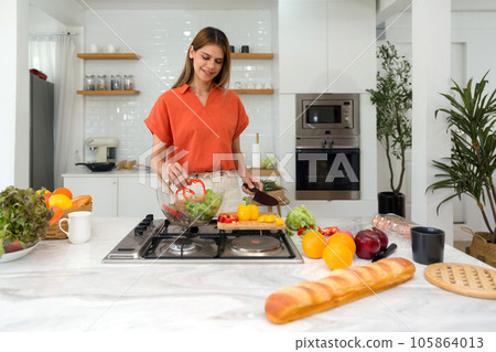 Young housewife enjoy making salad in a well-equipped, modern kitchen. The counter displays vibrant, fresh produce, revealing the beginnings of a nutritional meal. Young housewife enjoy making salad in a well-equipped, modern kitchen. The counter displays vibrant, fresh produce, revealing the beginnings of a nutritional meal. 105864013