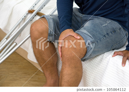 Injured man with painful knee resting on bed in an indoor setting, displaying discomfort. Crutches were placed beside him. 105864112
