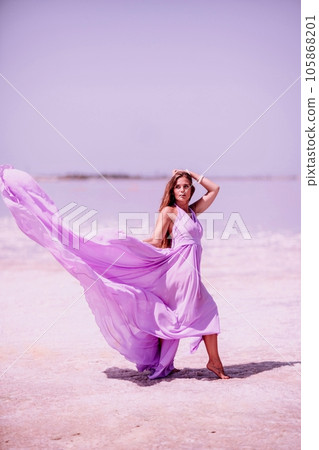 Woman pink salt lake. Against the backdrop of a pink salt lake, a woman in a long pink dress takes a leisurely stroll along the white, salty shore, capturing a wanderlust moment. 105868201